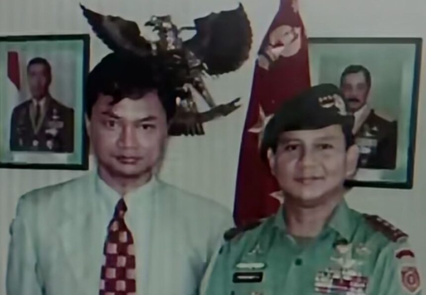 Two men posing for a photo: one in a light shirt and patterned tie, the other in a green military uniform with medals, with portraits and a flag in the background.