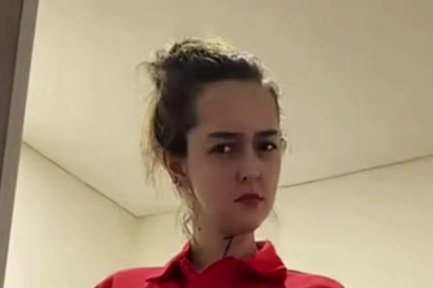 Close-up of a person with dark hair tied back, wearing a red shirt, looking to the side in an indoor setting.
